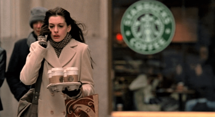 Anne Hathaway's character in the Devil Wears Prada carries 4 Starbucks coffee drinks in disposable cups on the street, with a Starbucks sign in the background