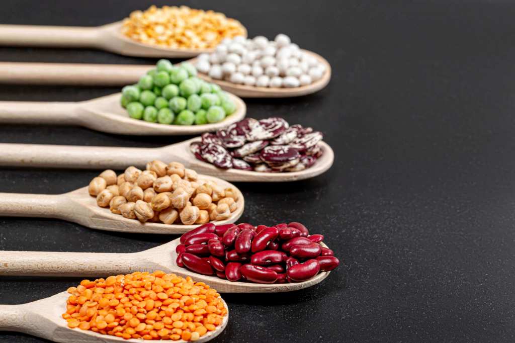 An assortment of legumes are displayed on wooden spoons against a black background.
