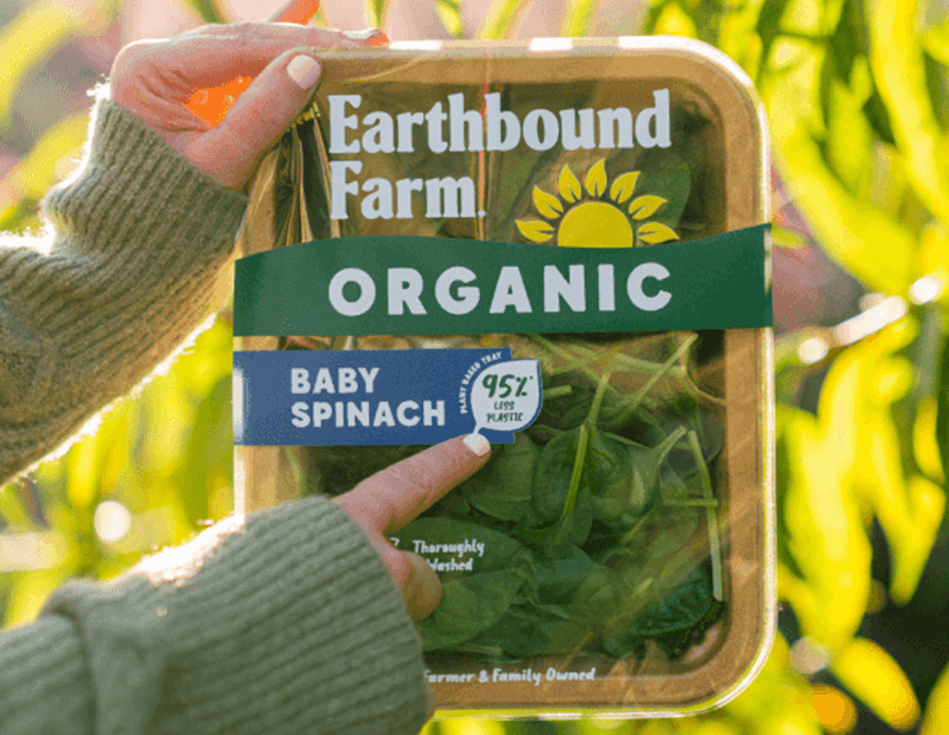 A picture of two hands holding up a box of Earthbound Farm organic baby spinach in the new compostable tray made from plant fibers. In the background are leaves in dappled light.