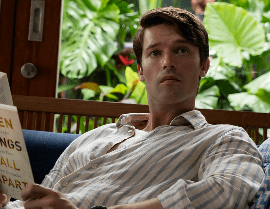 A screenshot from The White Lotus, Season 3. Saxon Ratliff (Patrick Schwarzenegger) sitting on a sofa in a resort reading When Things Fall Apart (Pema Chodron)