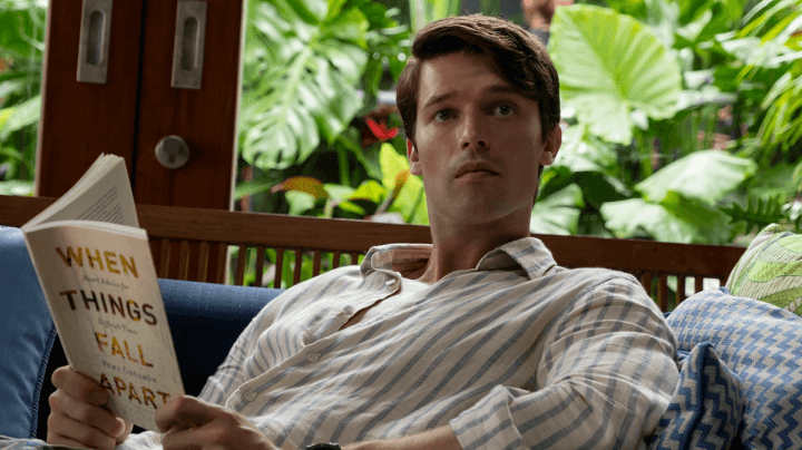 A screenshot from The White Lotus, Season 3. Saxon Ratliff (Patrick Schwarzenegger) sitting on a sofa in a resort reading When Things Fall Apart (Pema Chodron)