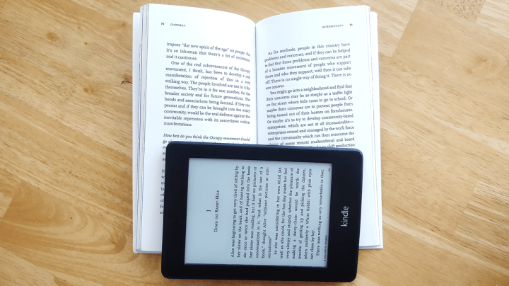 An Amazon Kindle lies on top of a physical book, keeping it open. 